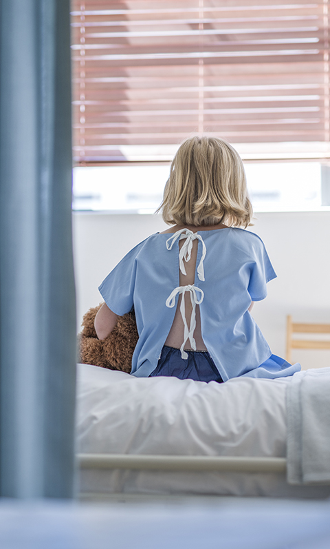 Rear view of sick girl sitting on hospital bed with curtain in the foreground